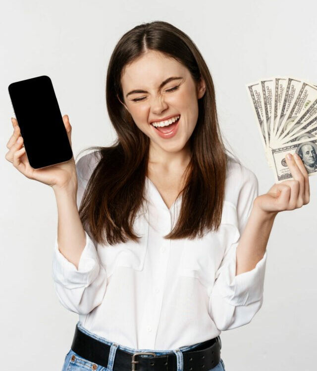 Enthusiastic young woman winning money, showing smartphone app interface and cash, microcredit, prize concept, standing over white background