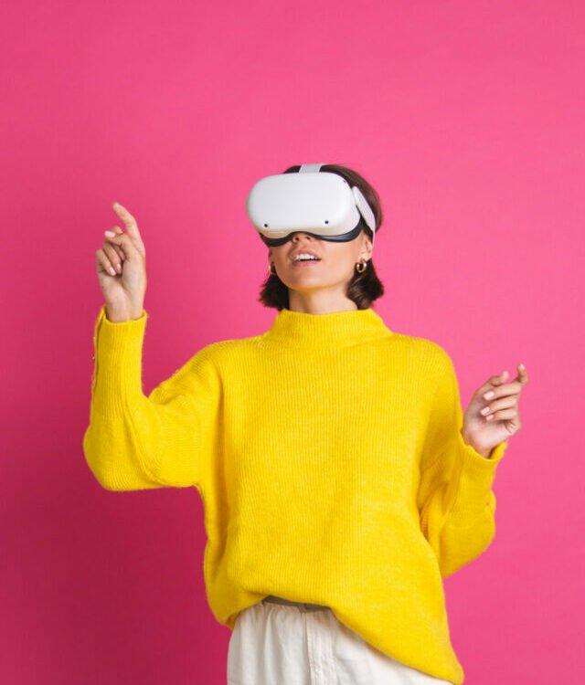Beautiful woman in bright yellow sweater on pink background in virtual reality glasses happy point left with finger to empty space